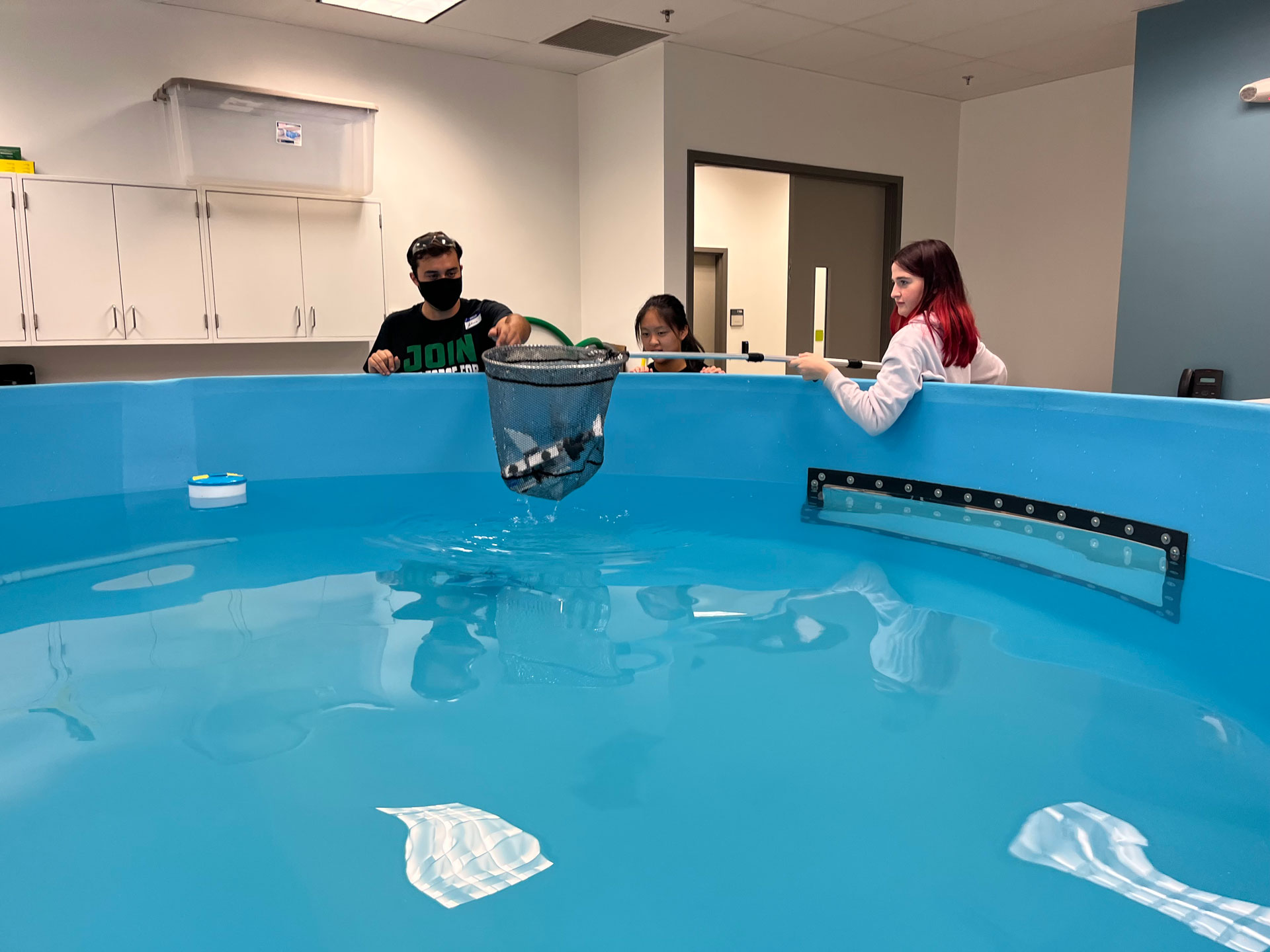 Three high school students use a net to gather their remotely operated vehicle out of a water tank