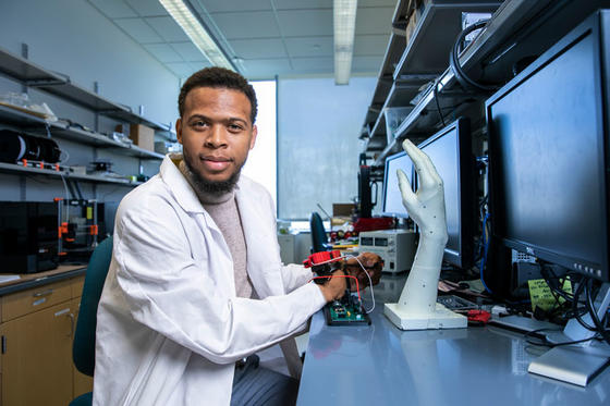 Quentin Sanders wears a white lab coat and beige turtleneck while operating a robotic arm in his lab.